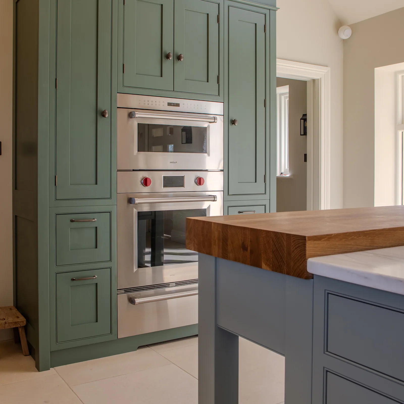 Built-in ovens set into green cabinetry beside a timber-topped island
