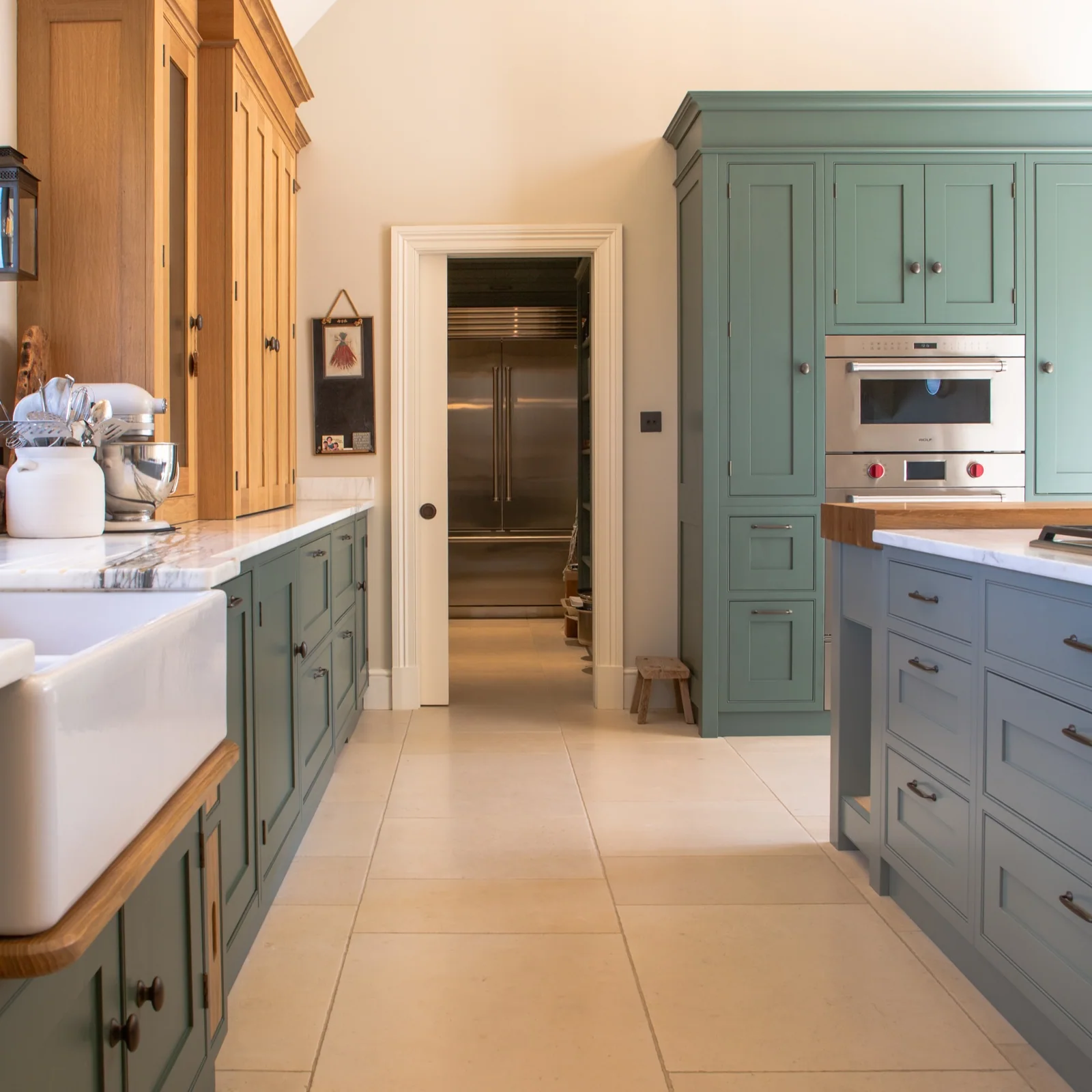 Green kitchen cabinetry with a white sink, built-in ovens and oak-framed windows
