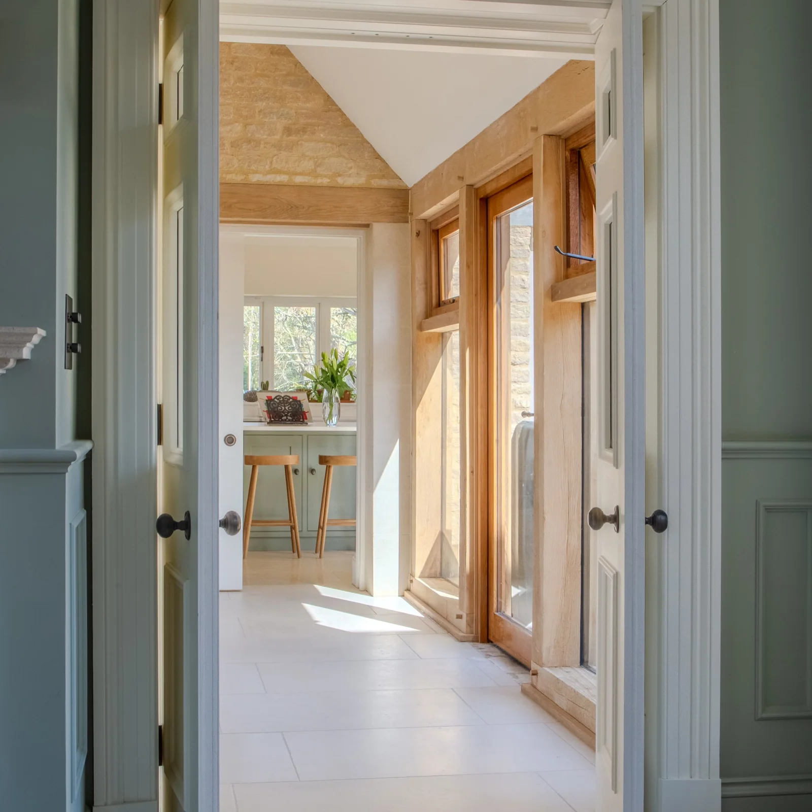 View through green kitchen cabinetry towards oak-framed glazing and a bright dining area
